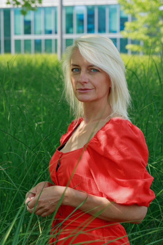 Portrait of a blonde woman in a red dress standing in tall grass outdoors.