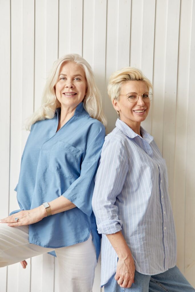 Two mature women in blue shirts smiling and posing indoors against a white wall.
