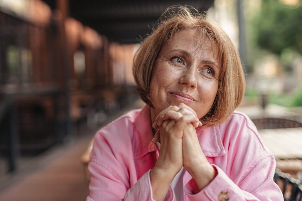 Senior woman with short brunette hair and pink shirt, hands clasped, looking thoughtful outdoors.